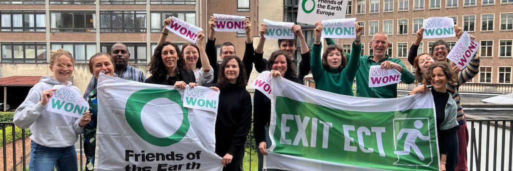 A group of people celebrating outdoor the EU Parliament voting forexiting the ECT,holding signis saying "we won!", a banner saying "Exit ECT" and flags with the Friends of the Earth Europe logo